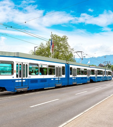 Ein altes weiss blaues Tram, das über eine Brücke fährt. Im Hintergrund sieht man einen Teil eines Sees und Teil der Stadt